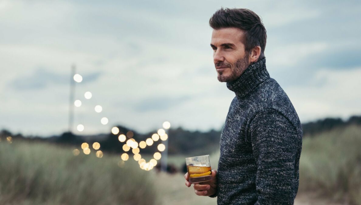 Porträtaufnahme von David Beckham in den Dünen mit einem Glas Whisky in seiner Hand