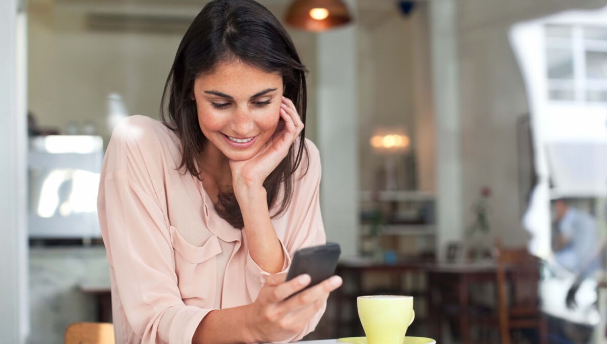 Eine junge Frau an einem Tisch in einem Café schaut lächelnd auf ihr Smartphone