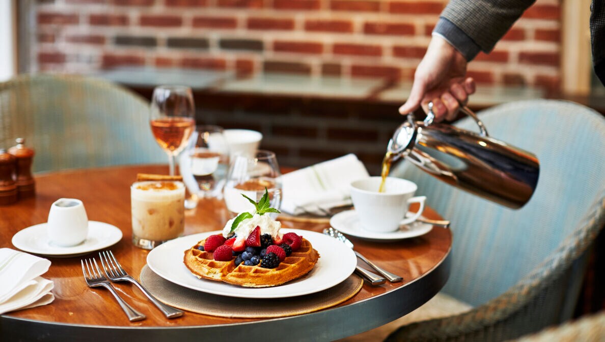 Eine Tasse Kaffee wird neben einem Teller mit Waffeln auf einem gedeckten Frühstückstisch eingegossen