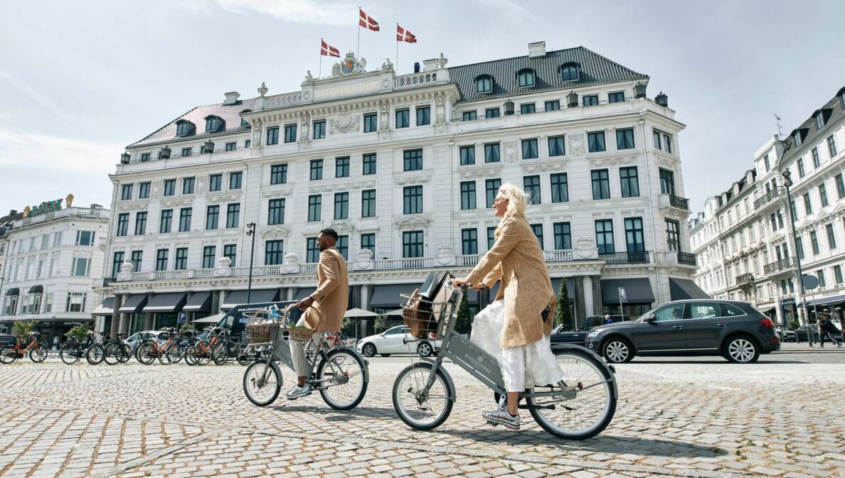 Zwei schick gekleidete Personen auf Fahrrädern vor dem neoklassizistischen Gebäude des Hotel d´Angleterre in Kopenhagen