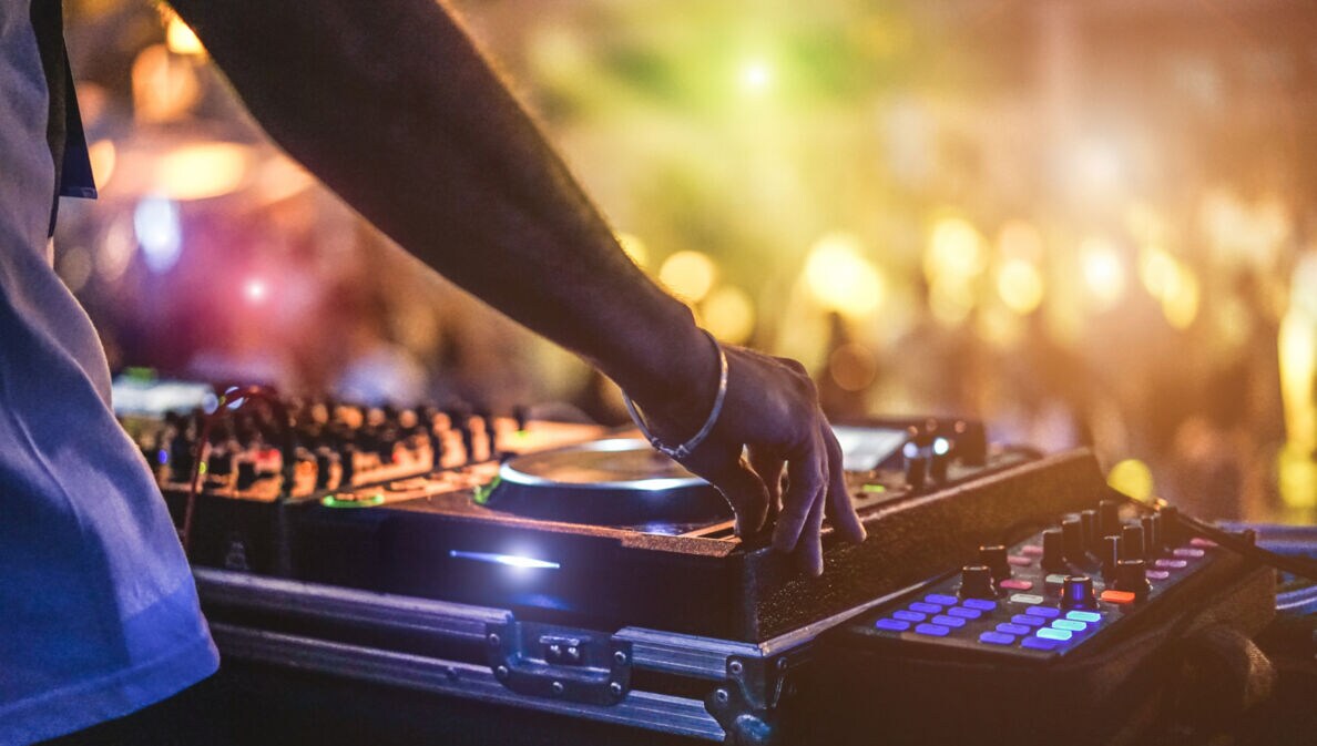 Die Hand eines DJs am Mischpult auf einem Festival mit Menschenmenge im Hintergrund
