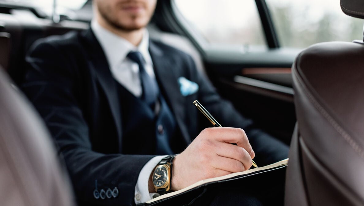 Ein Mann im Anzug sitzt in einem Auto und notiert etwas mit einem Kugelschreiber in einem Notizbuch