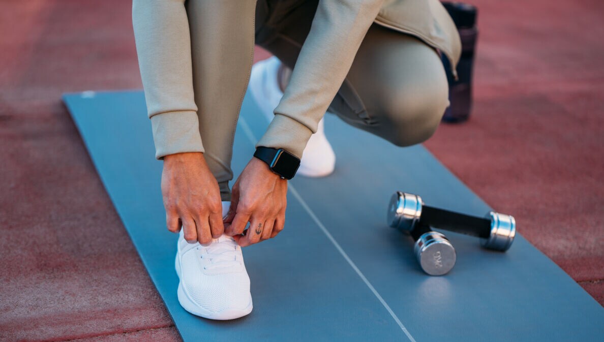 Eine Person in Sportkleidung mit Fitnessuhr am Handgelenk bindet sich die Schnürsenkel auf einer Sportmatte.