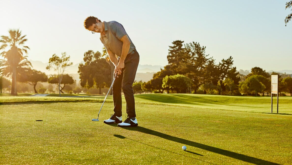 Ein Mann steht auf einem Golfplatzt und locht gerade den Golfball ein.