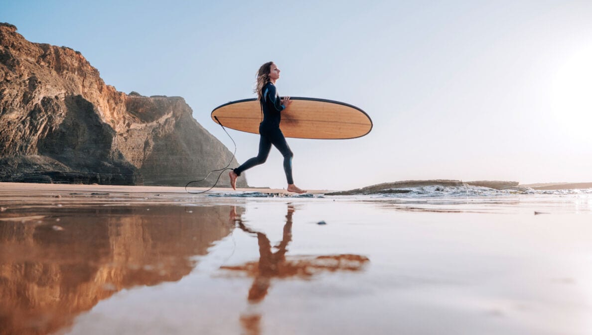 Eine Frau im Neoprenanzug trägt ihr Surfbrett unterm Arm ins Meer