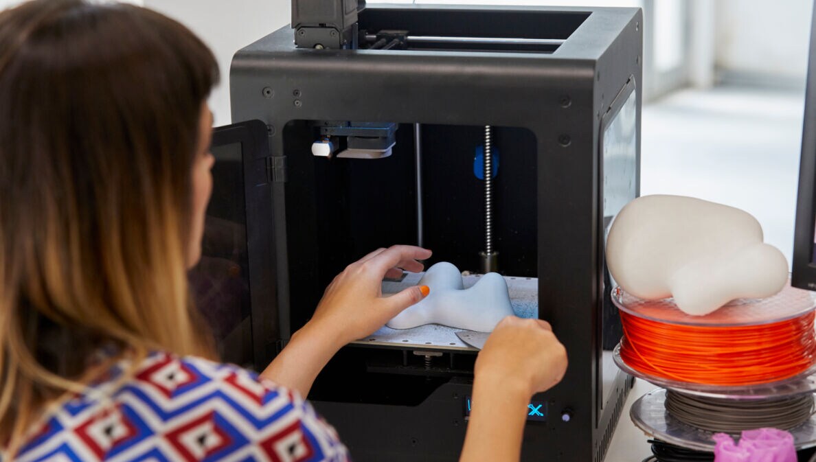 Rückansicht einer Frau, die ein kleines 3D-Objekt aus Kunststoff aus einem 3D-Drucker holt