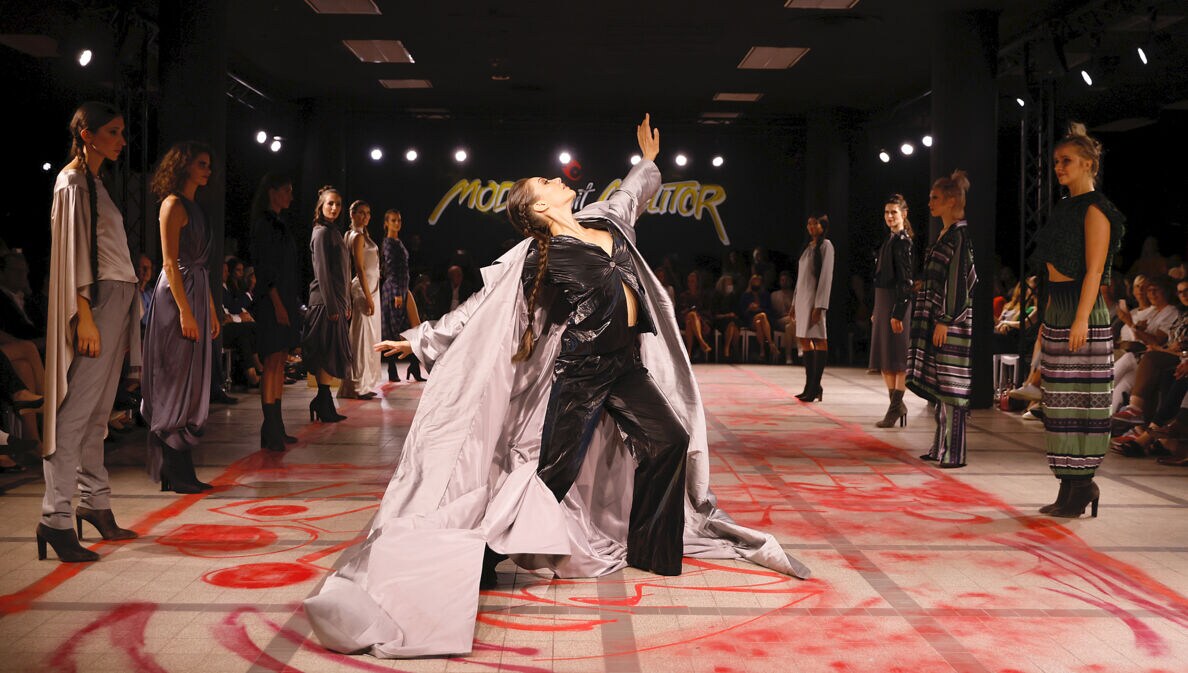 Eine Tänzerin mit weitem Mantel auf dem Catwalk umringt von stehenden Models