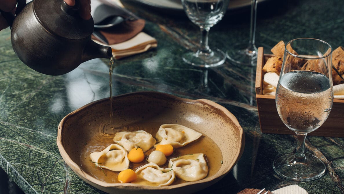 An einem stilvoll gedeckten Restauranttisch läuft Brühe aus einer Kanne in einen Teller mit gefüllter Pasta