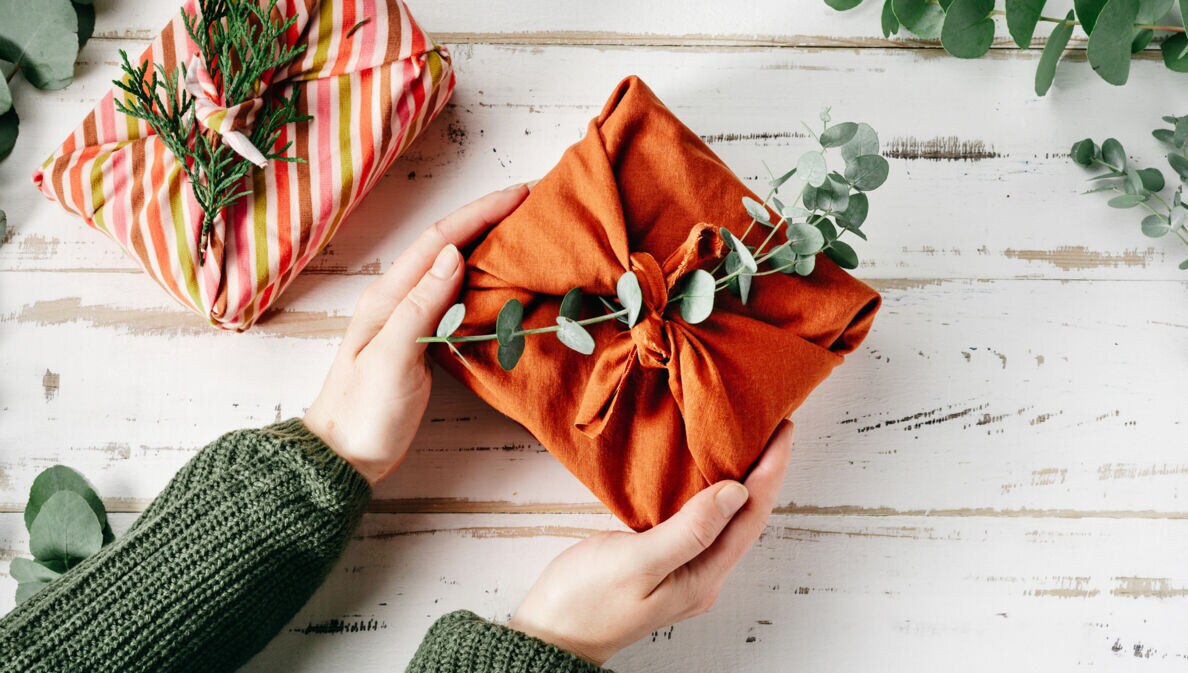 Ein Geschenk, verpackt in einem orangefarbenen Tuch und geschmückt mit einem grünen Zweig, wird von zwei Händen gehalten.