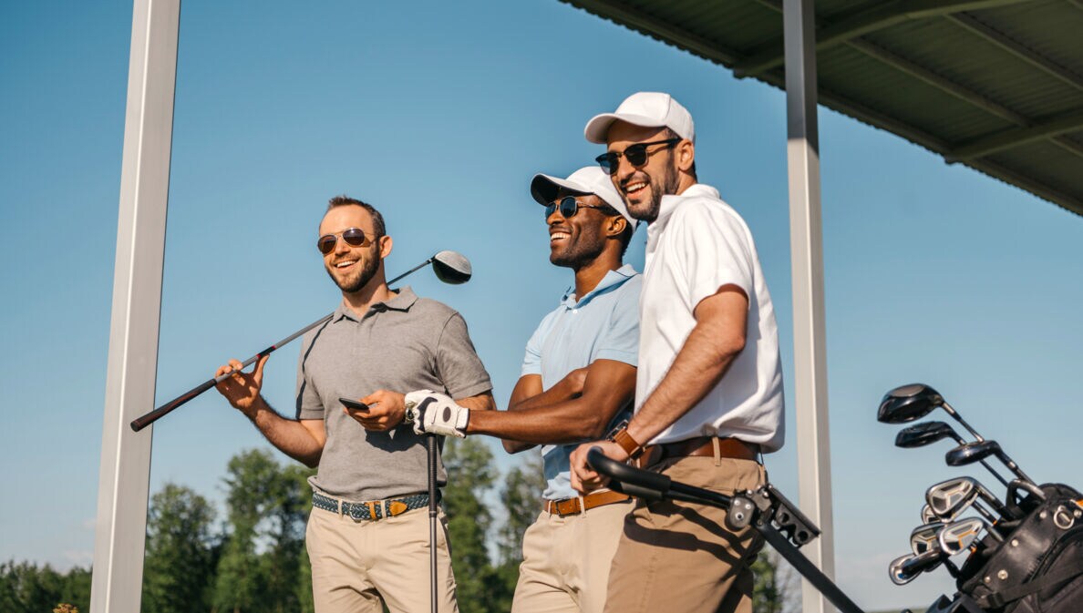 Drei Männer in Poloshirts, mit Sonnenbrillen und Golfschlägern stehen auf einer überdachten Terrasse und lachen