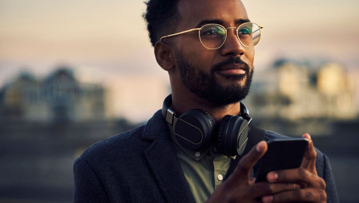 Ein junger Mann mit Kopfhörern um den Hals hält ein Smartphone in der Hand und schaut in die Ferne