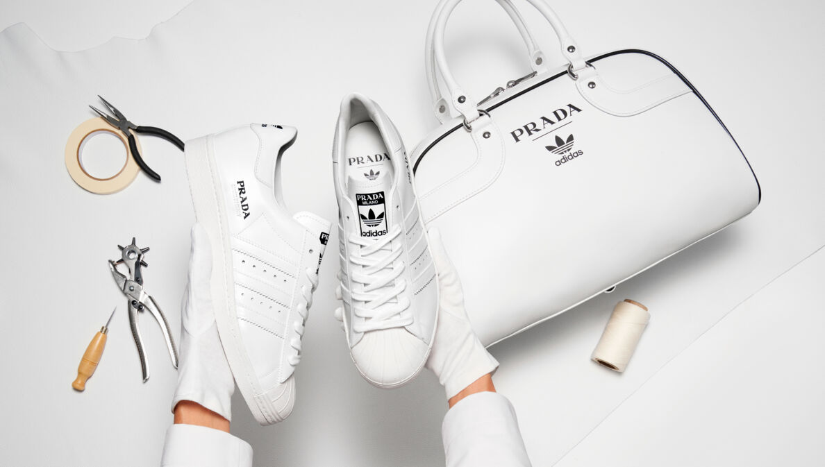 Weiße Turnschuhe neben einer Handtasche mit Logos der Marken adidas und Prada auf weißem Untergrund