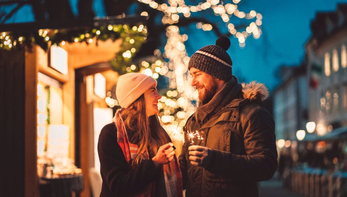 Eine Frau und ein Mann stehen auf dem Weihnachtsmarkt und schauen sich an, in ihren Händen halten sie Wunderkerzen