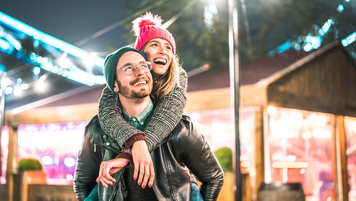 Ein Mann trägt eine Frau in weihnachtlicher Atmosphäre Huckepack, beide lächeln dabei