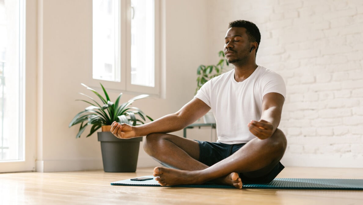 Ein Mann sitzt im Schneidersitz auf einer Yogamatte und meditiert mit einer Meditations-App