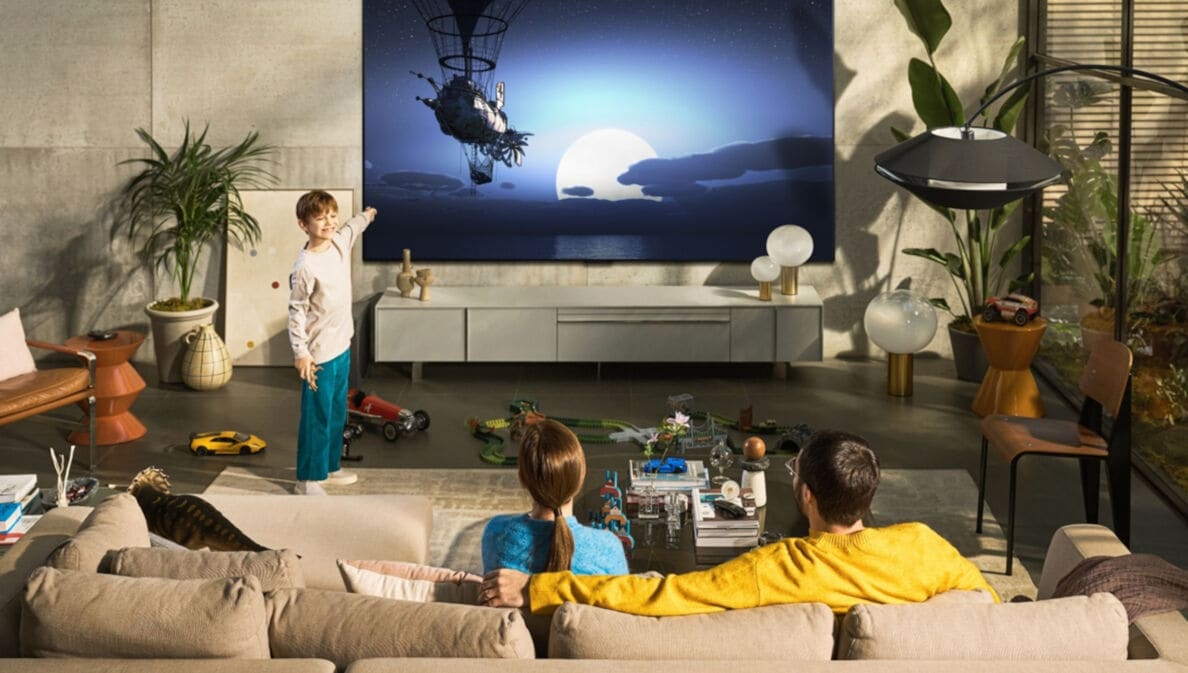 Ein Mann und eine Frau sitzen auf einem beigen Sofa und schauen auf einen großen Fernseher, davor steht ein Junge und zeigt auf den Bildschirm