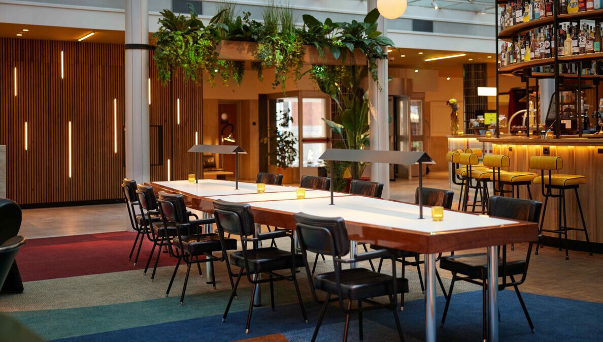 Langer Tisch mit Stühlen in einer Hotel-Lobby, im Hintergrund eine beleuchtete Bar