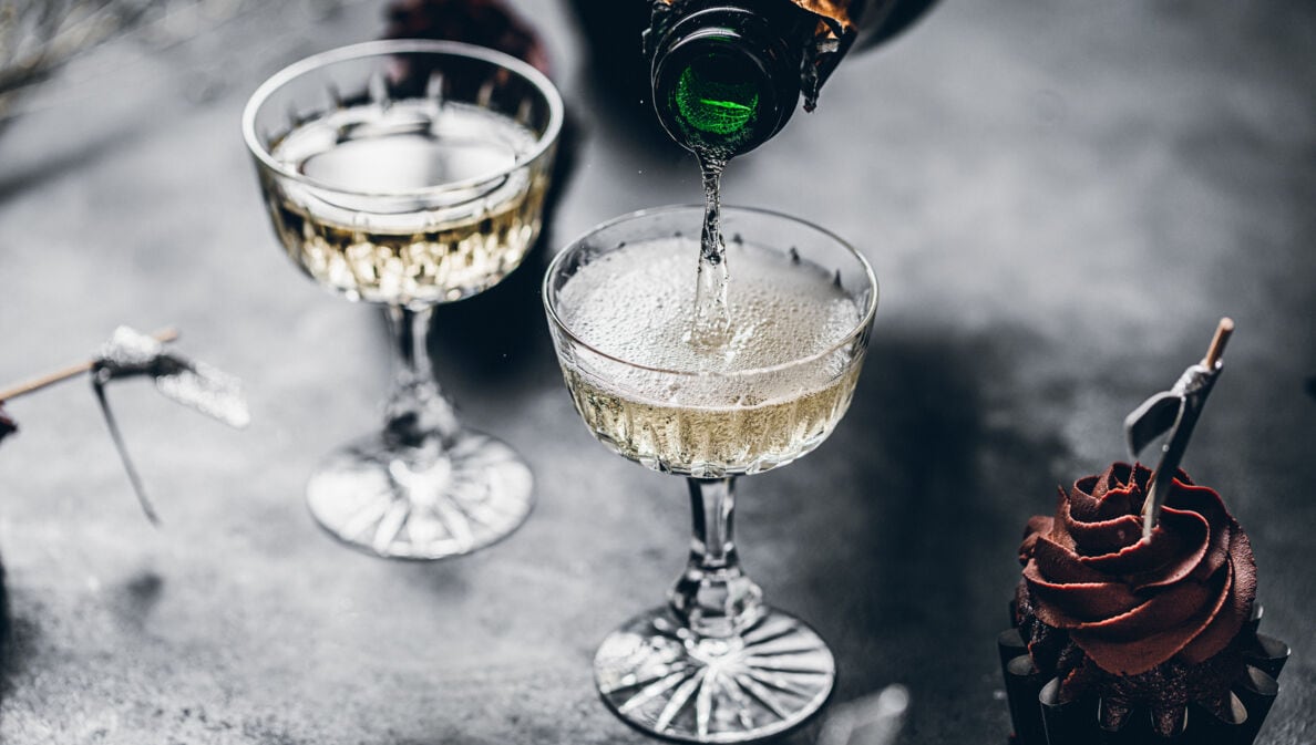 Zwei Gläser auf einem grauen Tisch werden mit Champagner gefüllt, daneben ist ein Cupcake mit Schokohaube zu sehen