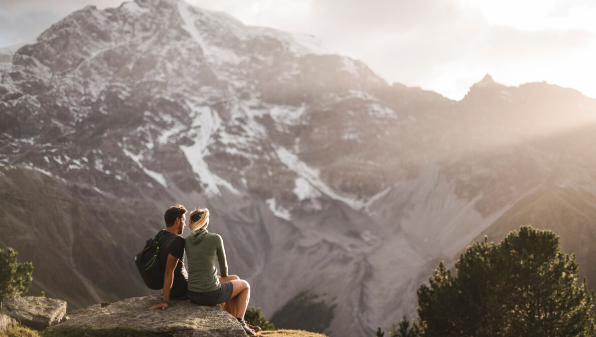 Zwei Personen sitzen im Nationalpark Stilfserjoch auf einem Stein und blicken auf ein Bergmassiv.