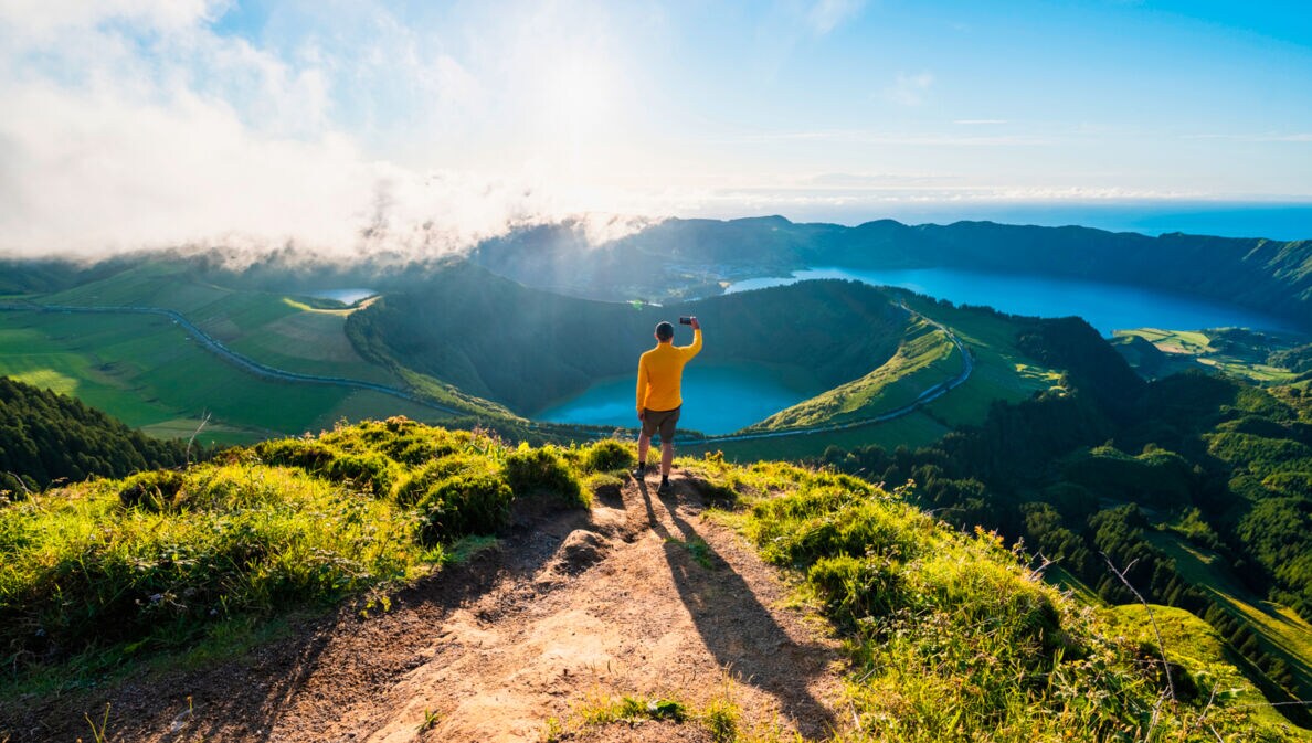 Eine Person im gelben Oberteil steht auf einem Berg und blickt auf einen Vulkan auf den Azoren