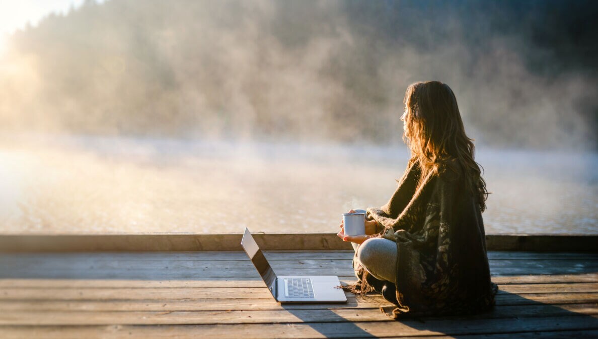 Eine Frau sitzt entspannt mit einer Kaffeetasse vor einem Laptop an einem nebelverhangenen See am Morgen