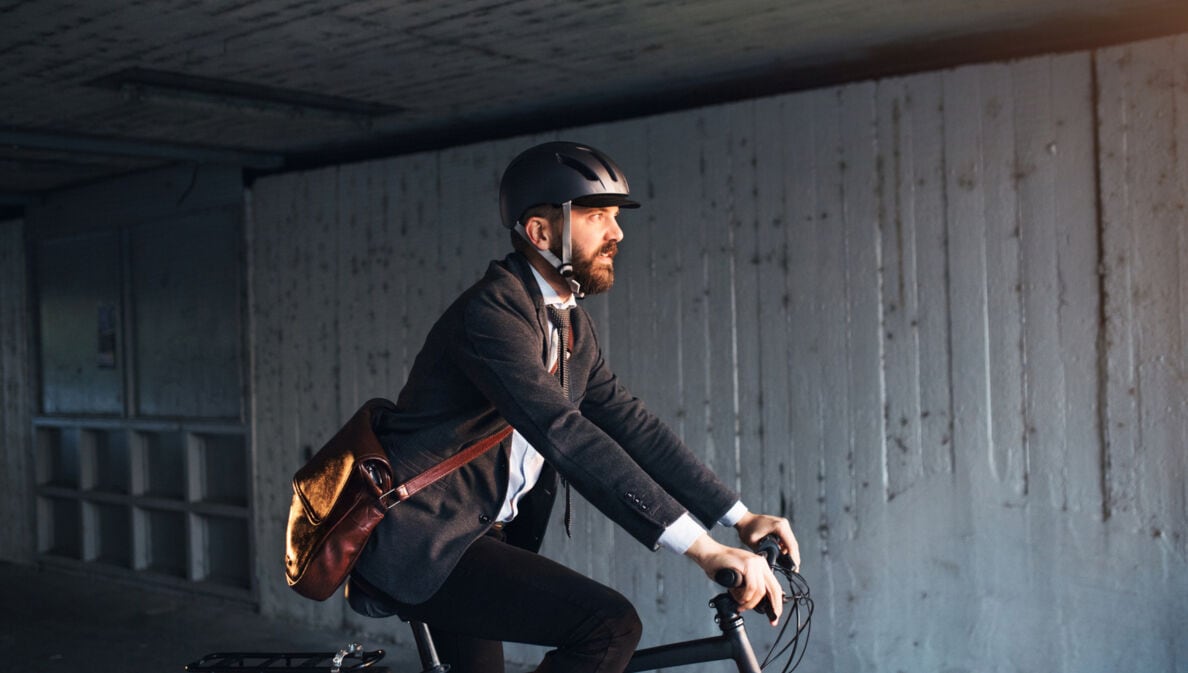 Seitenansicht einer Person im Anzug, mit Umhängetasche und Helm, die auf einem Fahrrad fährt