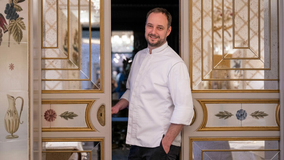 Der Koch Franz-Josef Unterlechner steht in einer geöffneten Flügeltür mit goldenen Verzierungen, Malereien und Spiegelelementen