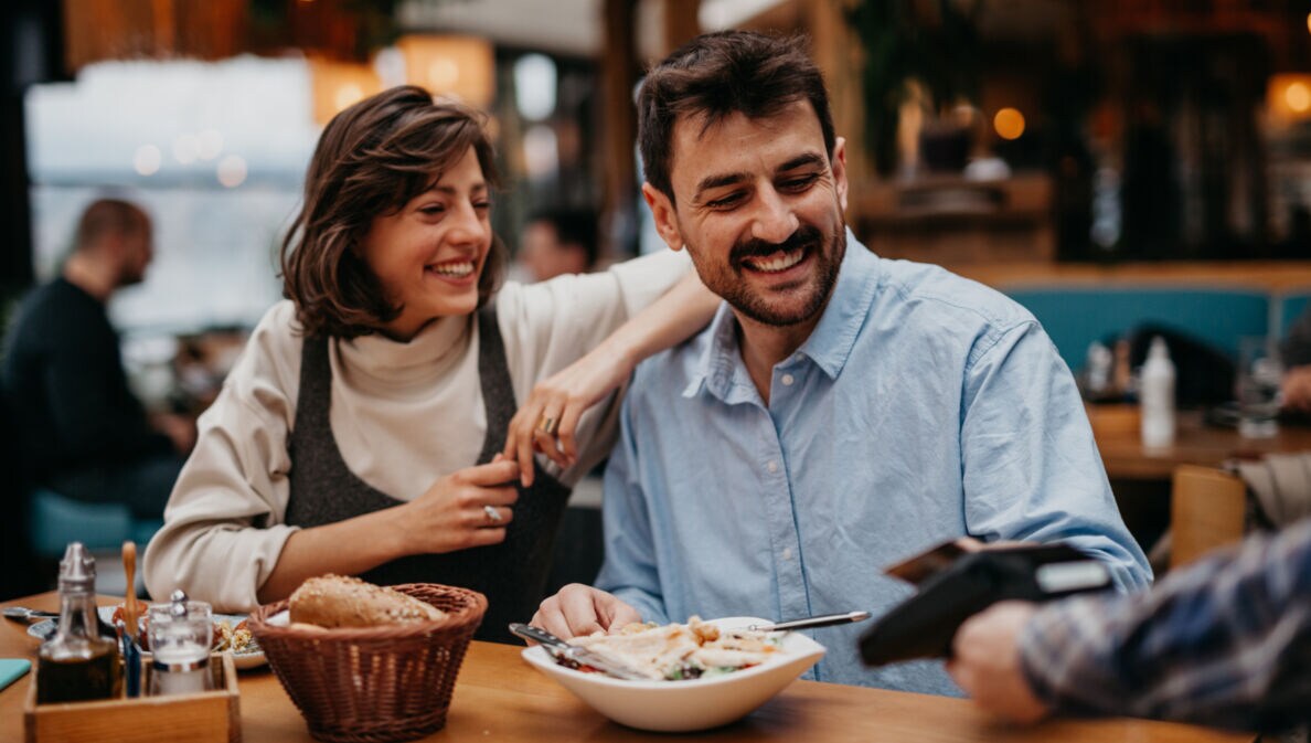 Eine Frau mit braunen Haaren und ein Mann mit Bart sitzen im Restaurant und lächeln, während er dem Kellner seine Kreditkarte zum bezahlen reicht
