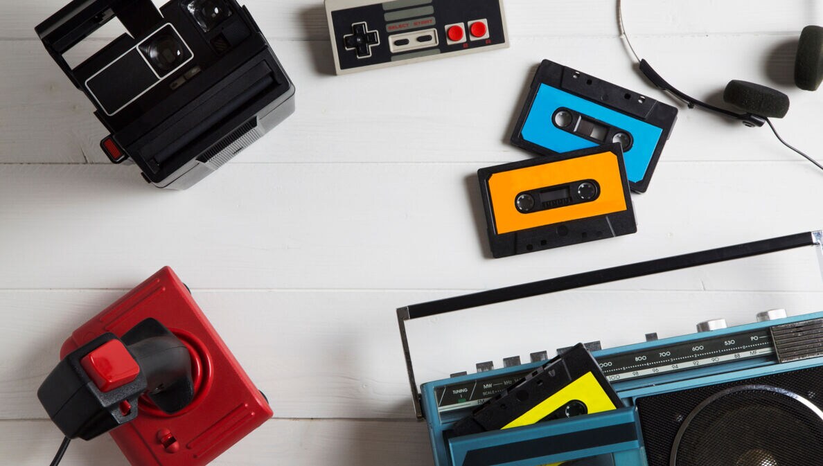 Auf einem Tisch liegen mehrere analoge Medien in Form von Kassetten, einer Polaroidkamera, einem Kassettenrekorder, einem NES-Gamepad und Joystick