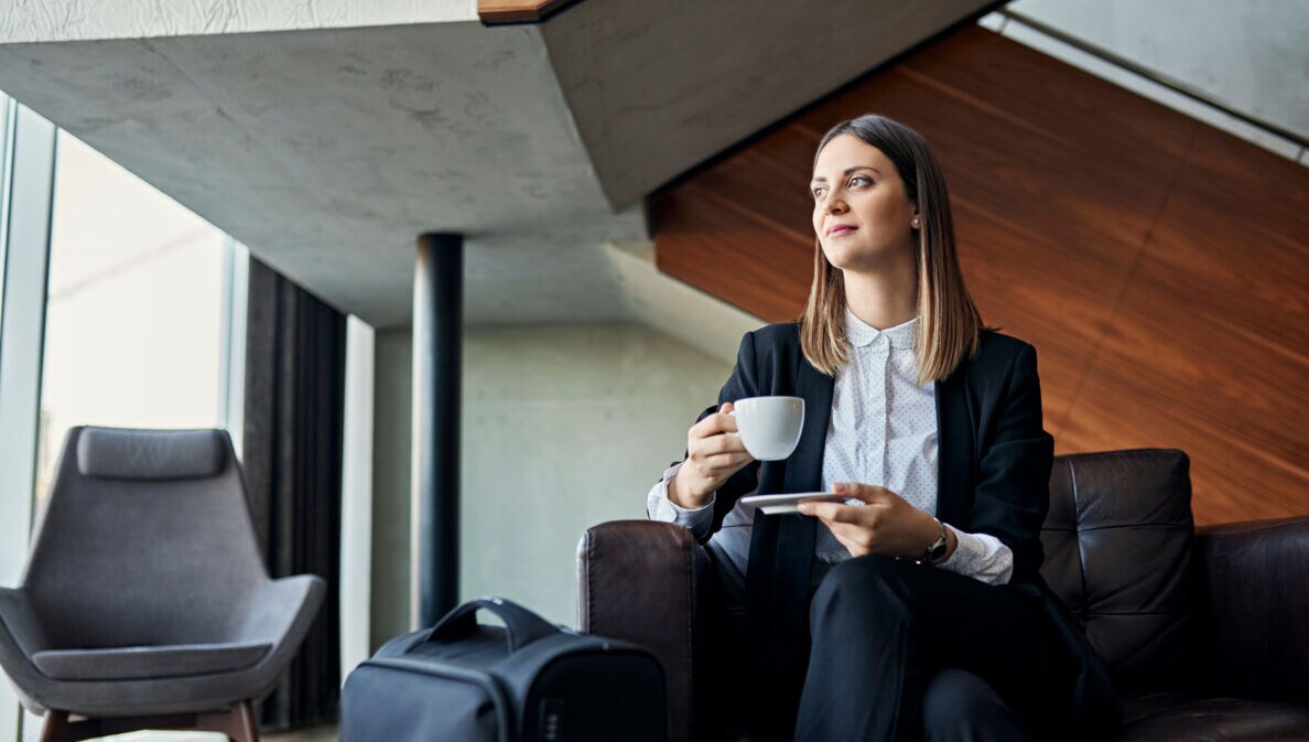 Eine junge Geschäftsfrau genießt die Wartezeit mit einer Tasse Kaffee auf einem Sessel in einer Lounge