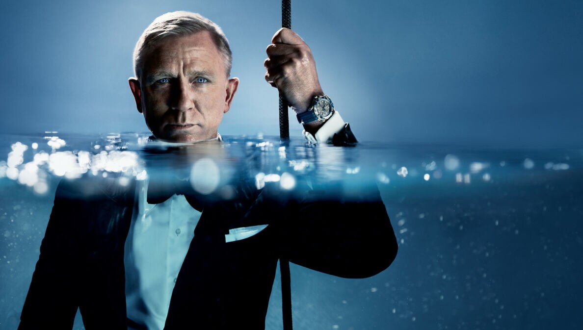 Porträt von Daniel Craig im schwarzen Smoking im Wasser, das ihm bis zum Hals steht; an seiner hochgehaltenen Hand trägt er eine Taucheruhr der Marke OMEGA