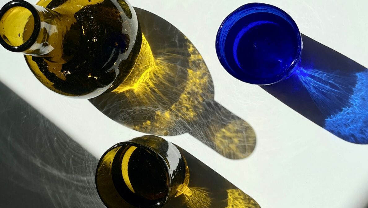 Eine olivgrüne Glaskaraffe, ein olivgrünes und ein blaues Glas, durch die von einer Seite fällt Licht fällt, wodurch farbliche Schatten entstehen.