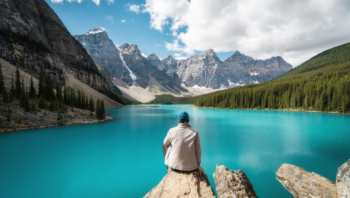 Eine Person sitzt auf einem Stein und schaut auf einen See, der gesäumt von bewaldeten Bergen ist