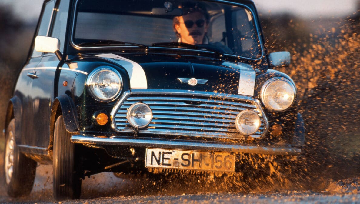 Ein Mann fährt in einem schwarzen MINI Cooper 1.3 mit Rallystreifen durch eine Schlammpfütze