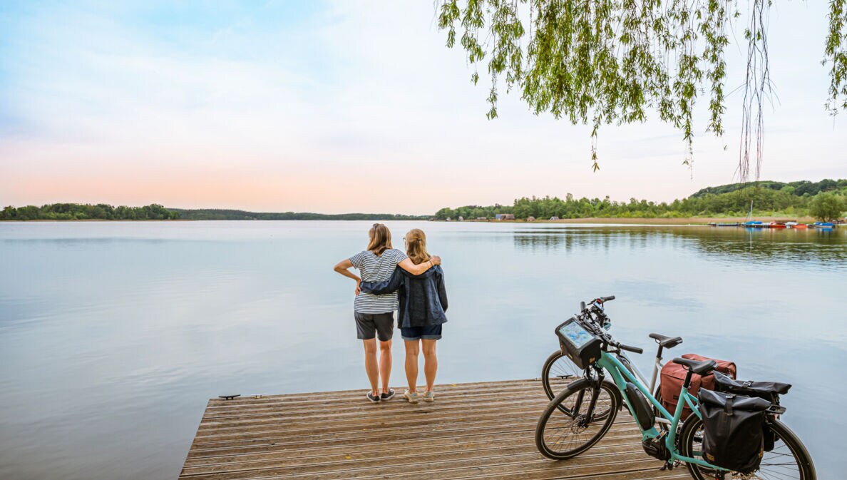 Rückansicht von zwei Frauen, die auf einem Steg vor einem See stehen und sich umarmen, daneben zwei E-Bikes
