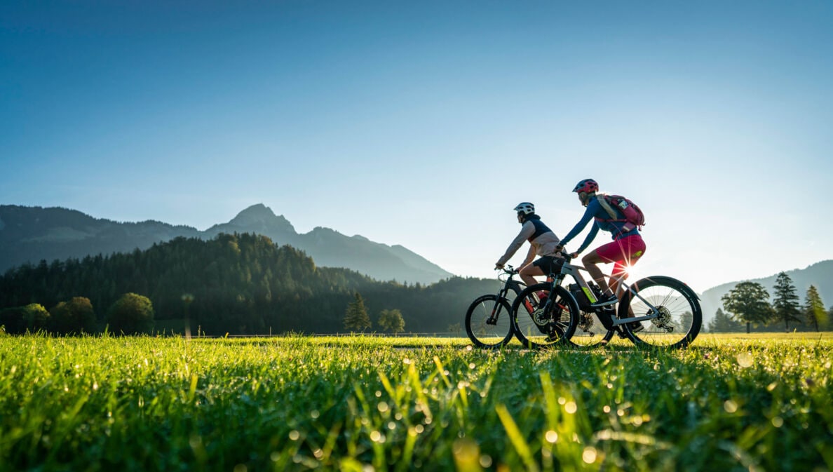 Zwei Personen fahren auf Mountainbikes durch eine Grasebene vor Bergpanorama im Sonnenschein