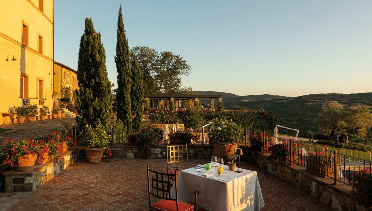 Ein gedeckter Tisch auf einer großen Terrasse einer Villa im toskanischen Baustil mit Blick in eine grüne Hügellandschaft bei Sonnenuntergang
