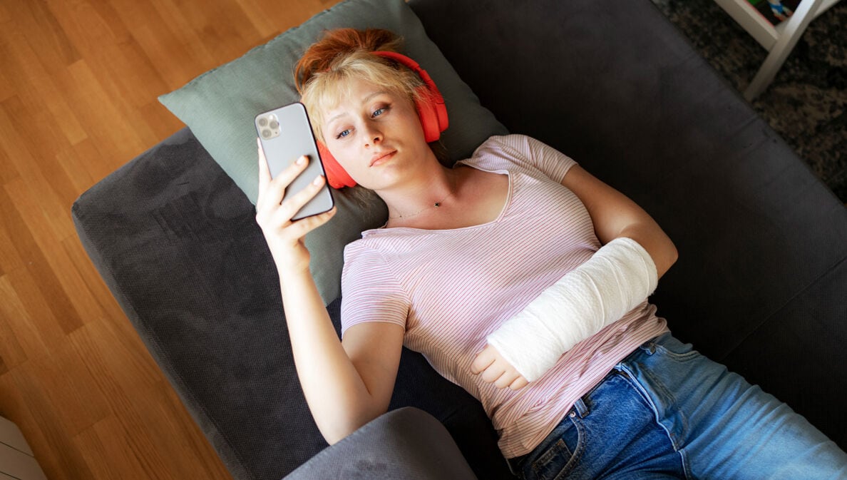 Aufsicht einer auf einem Sofa liegenden, jungen Frau, die mit Kopfhörern und einem eingegipsten Arm auf ihr Smartphone schaut.