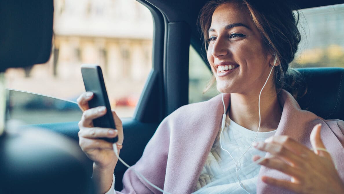 Eine junge Frau mit Kopfhörern auf dem Rücksitz eines Autos schaut lachend auf das Display ihres Smartphones.