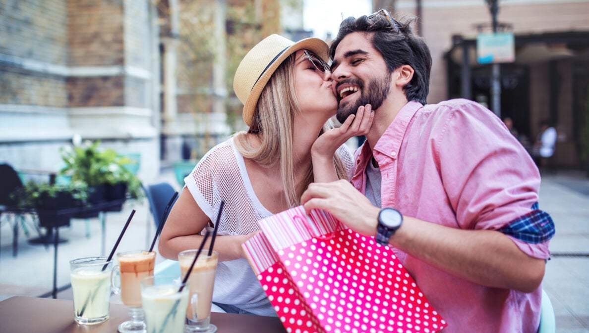 Ein junges, glückliches Paar mit einer roten Geschenktüte an einem Tisch im Außenbereich eines Cafés, sie gibt ihm einen Kuss auf die Wange.