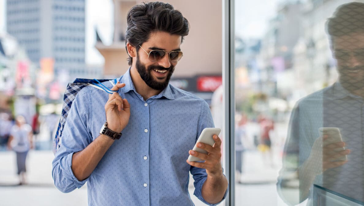 Ein junger Mann mit Vollbart, Sonnenbrille und Einkaufstüten in der Hand schaut in einer Einkaufsstraße lächelnd auf sein Smartphone.