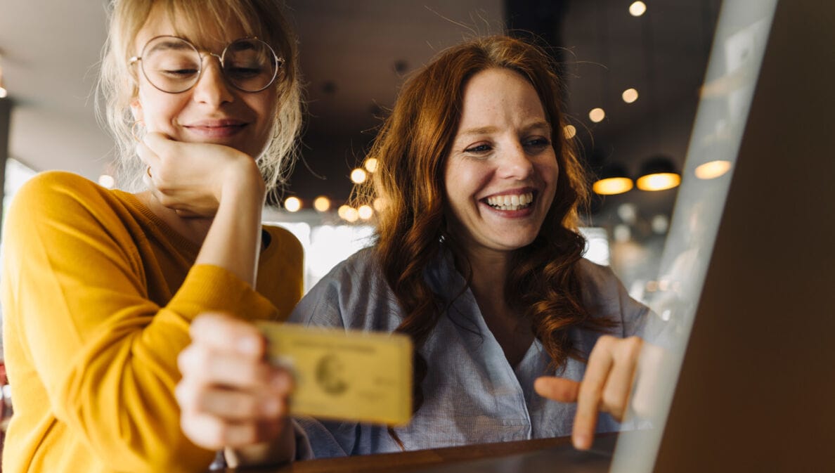 Zwei junge Frauen sitzen lächelnd mit einer Kreditkarte vor einem Laptop in einem Café.