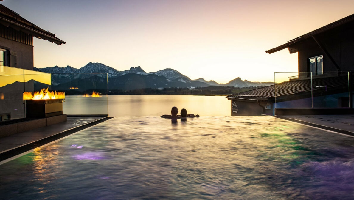 Zwei Personen in einem Infinity-Pool schauen über einen See in eine Berglandschaft bei Sonnenuntergang
