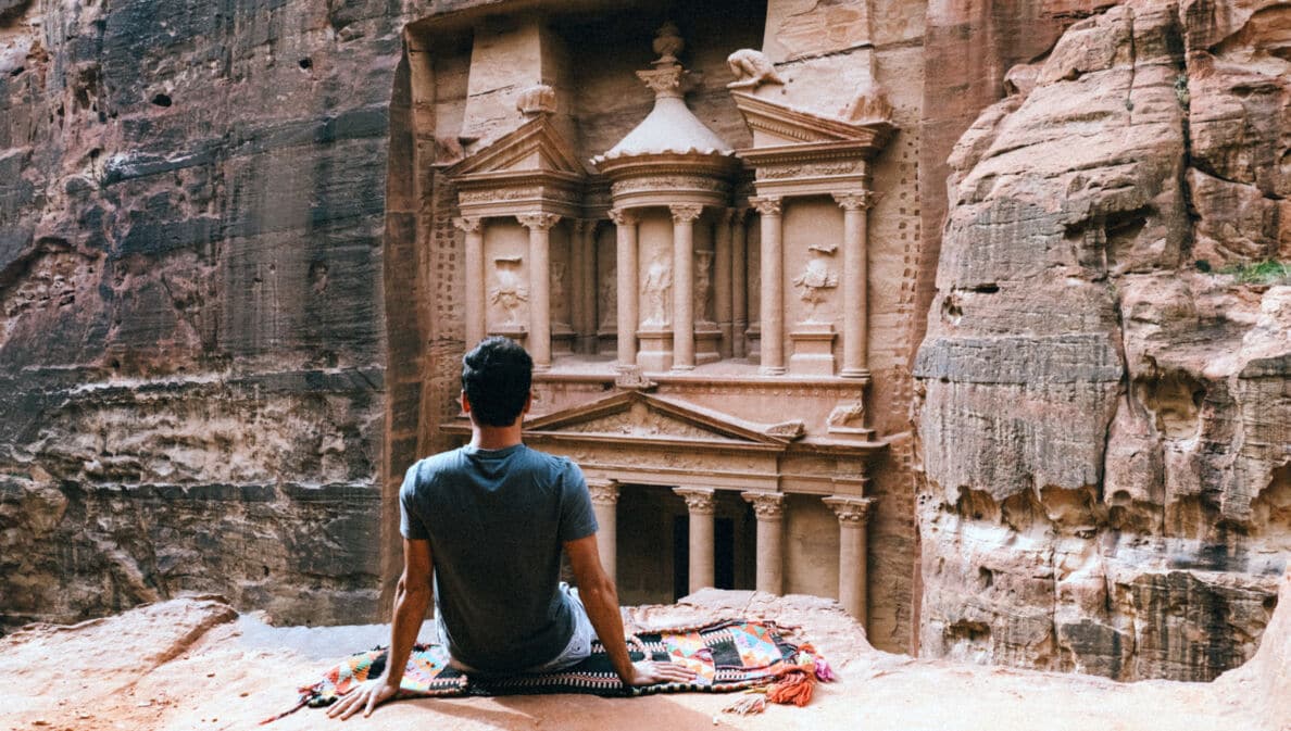 Rückansicht eines auf einem Felsvorsprung sitzenden Mannes vor dem Eingang eines monumentalen Grabtempels in der Felsenstadt Petra