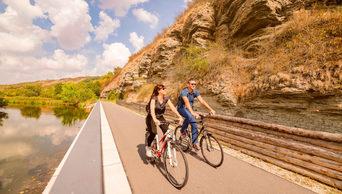Zwei Personen fahren auf Fahrrädern auf einem asphaltierten Radweg vor einer Steinwand an einem Fluss entlang