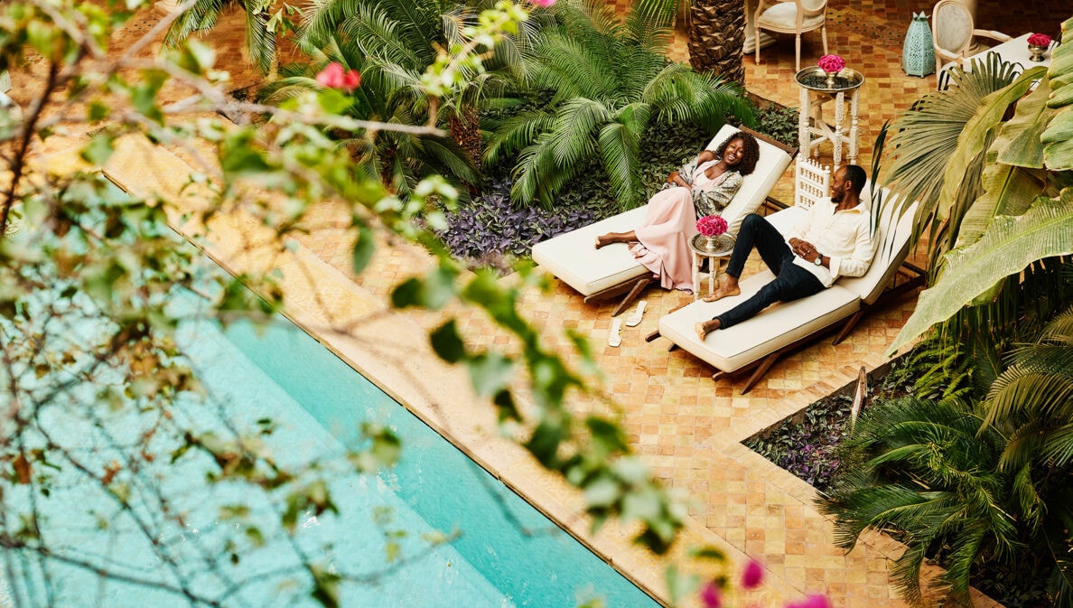 Aufsicht eines elegant gekleideten Paares auf Sonnenliegen an einem Pool auf der Terrasse eines Boutiquehotels.