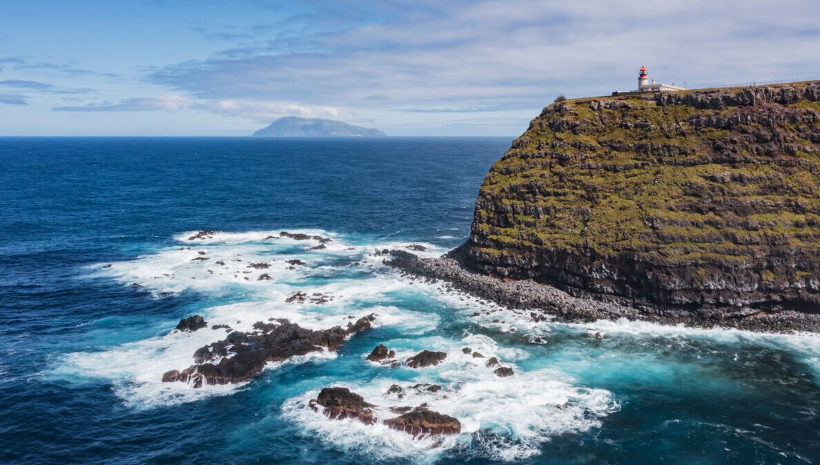 Luftaufnahme einer Steilklippe mit Leuchtturm und der Küste vorgelagerte Felsen im Meer, im Hintergrund eine Insel