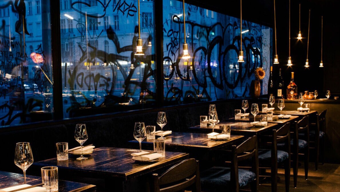 Beleuchtete, eingedeckte Holztische in einem modernen Restaurant vor einer Fensterfront mit Graffiti bei Nacht.