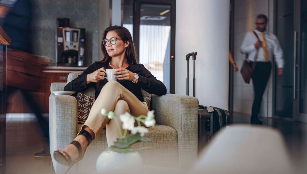 Eine Frau sitzt mit einer Kaffeetasse in einem Sessel in einer belebten Lounge, neben ihr ein Reisekoffer.