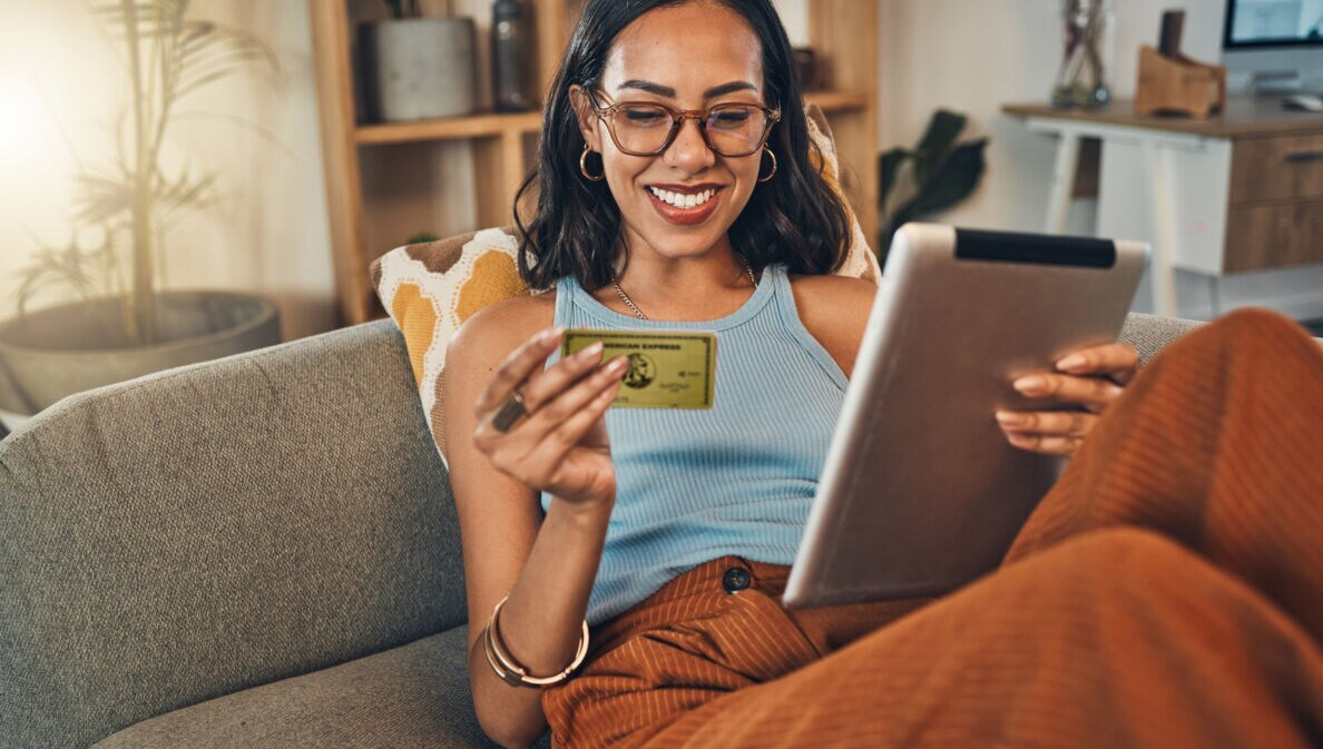 Eine junge Frau sitzt lächelnd auf einem Sofa, in ihren Händen hält sie ein Tablet und eine goldene Kreditkarte von American Express.